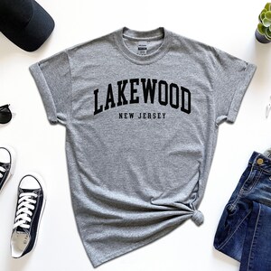 May include: A heather gray t-shirt with the word "Lakewood" arched across the chest, with "New Jersey" below. The shirt is styled with black sunglasses, black sneakers, a black cap, a smartwatch, and blue jeans. A small succulent plant is also present.