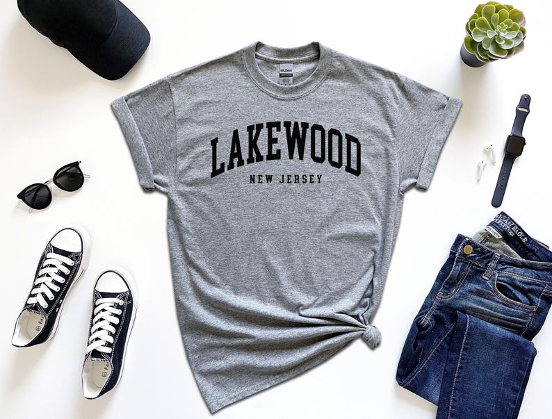 May include: A heather gray t-shirt with the word "Lakewood" arched across the chest, with "New Jersey" below. The shirt is styled with black sunglasses, black sneakers, a black cap, a smartwatch, and blue jeans. A small succulent plant is also present.