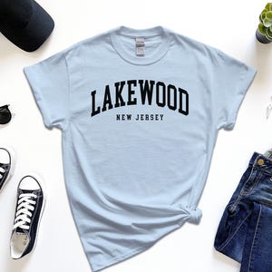 May include: A light blue t-shirt with the word "LAKEWOOD" arched across the chest, with "NEW JERSEY" below in black text. The shirt is styled with black sunglasses, sneakers, a watch, a cap, and jeans.