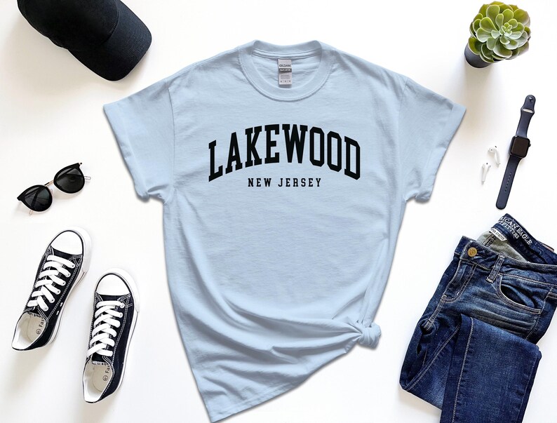 May include: A light blue t-shirt with the word "LAKEWOOD" arched across the chest, with "NEW JERSEY" below in black text. The shirt is styled with black sunglasses, sneakers, a watch, a cap, and jeans.