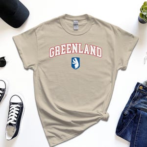 Op de afbeelding: Een beige t-shirt met een rode en blauwe afbeelding met de tekst "GREENLAND" met een witte ijsbeer in een blauw schild.