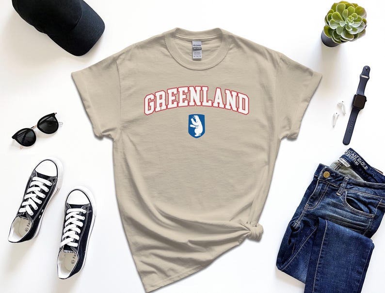 Op de afbeelding: Een beige t-shirt met een rode en blauwe afbeelding met de tekst "GREENLAND" met een witte ijsbeer in een blauw schild.