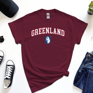 Op de afbeelding: Een maroon t-shirt met witte tekst die "GREENLAND" zegt en een blauwe en witte afbeelding van een ijsbeer.
