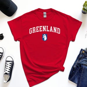 Op de afbeelding: Rood T-shirt met witte tekst "GREENLAND" en een blauwe en witte afbeelding van een ijsbeer.