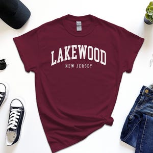 May include: A maroon t-shirt with the text "Lakewood New Jersey" in white, paired with black sunglasses, black sneakers, a black cap, a watch, and blue jeans. A small succulent plant is also visible.