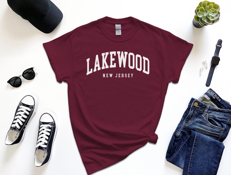 May include: A maroon t-shirt with the text "Lakewood New Jersey" in white, paired with black sunglasses, black sneakers, a black cap, a watch, and blue jeans. A small succulent plant is also visible.