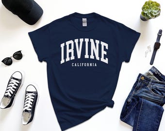 May include: Navy blue t-shirt with the word "IRVINE" in white block letters above "CALIFORNIA". The shirt is surrounded by black sunglasses, black sneakers, a black cap, a watch, and blue jeans.