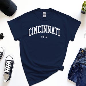 Puede incluir: Camiseta azul marino con la palabra "CINCINNATI" en letras blancas arqueadas en el pecho, con "OHIO" debajo. La imagen incluye gafas de sol negras, zapatillas deportivas, un reloj y vaqueros.