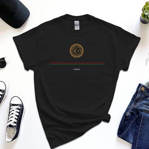Könnte beinhalten: Schwarzes T-Shirt mit einem goldenen Emblem, das einen Halbmond und einen Stern zeigt, mit dem Wort "LIBYA" darunter gedruckt. Das Shirt liegt flach auf einer weißen Oberfläche mit einer Jeans, einer Sonnenbrille und Sneakers.