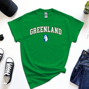 Op de afbeelding: Groen T-shirt met het woord "GREENLAND" in witte letters met een rode omtrek en een witte ijsbeergrafiek op een blauwe achtergrond.