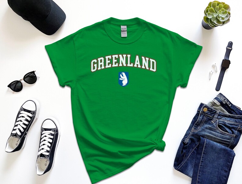 Op de afbeelding: Groen T-shirt met het woord "GREENLAND" in witte letters met een rode omtrek en een witte ijsbeergrafiek op een blauwe achtergrond.