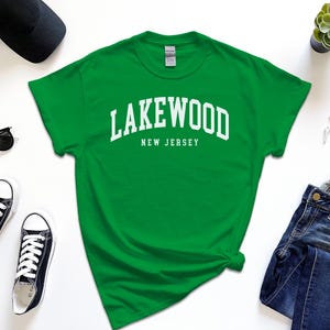May include: A green t-shirt with the text "Lakewood New Jersey" in white block letters. The shirt is surrounded by a black baseball cap, sunglasses, black sneakers, a succulent plant, a smartwatch, and blue jeans.