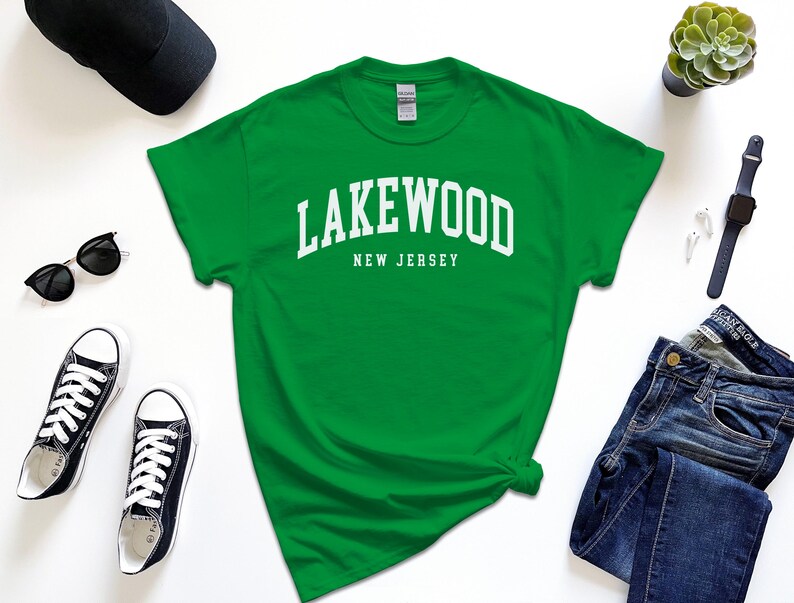 May include: A green t-shirt with the text "Lakewood New Jersey" in white block letters. The shirt is surrounded by a black baseball cap, sunglasses, black sneakers, a succulent plant, a smartwatch, and blue jeans.