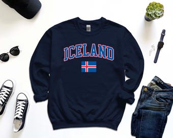 Puede incluir: Sudadera azul marino con la palabra "ICELAND" en blanco con contorno rojo y la bandera islandesa. La sudadera es de cuello redondo. La imagen también incluye gafas de sol negras, zapatillas negras y unos vaqueros azules.