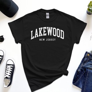 May include: Black t-shirt with the white text "Lakewood New Jersey". The shirt is surrounded by black sunglasses, a black baseball cap, black sneakers, blue jeans, a watch, and a small succulent plant.