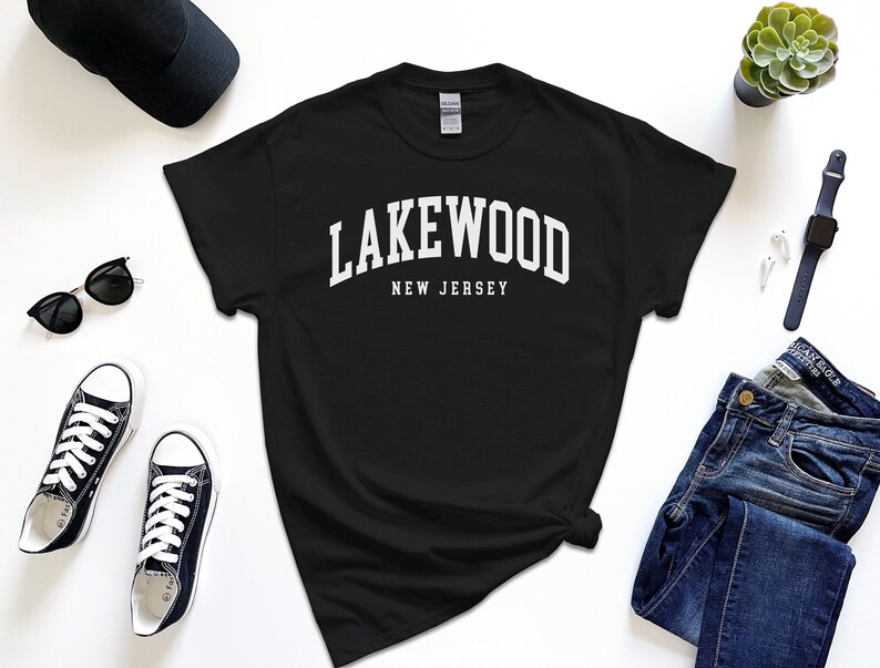 May include: Black t-shirt with the white text "Lakewood New Jersey". The shirt is surrounded by black sunglasses, a black baseball cap, black sneakers, blue jeans, a watch, and a small succulent plant.