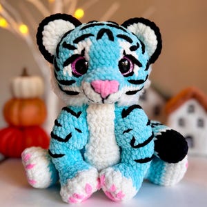 May include: A blue and white crocheted tiger plush toy with pink paw pads and black stripes. The toy has large black eyes with pink glitter.