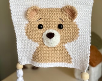 May include: A white crocheted wall hanging with a brown bear face in the centre. The bear has black eyes and a brown nose. The hanging has two white tassels with wooden beads at the top.