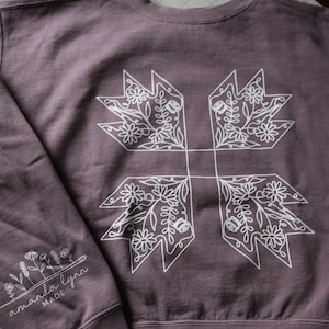 May include: A mauve long-sleeve crewneck sweatshirt featuring a large white line-art graphic of a four-pointed floral quilt block on the chest. The left sleeve displays a smaller white floral illustration with the text amanda lynn MADE.