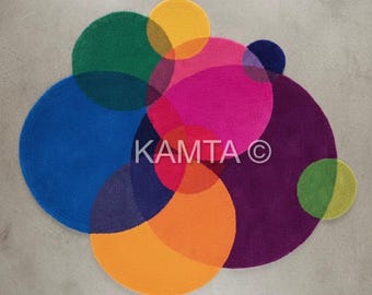 May include: A colorful rug featuring overlapping circles in shades of blue, green, yellow, pink, orange, and purple. The design includes the text "KAMTA ©". The rug has a soft, textured appearance.
