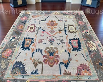 May include: A large, rectangular area rug with an intricate, colorful pattern. The rug features a light gray central field with a border of darker gray and accents of blue, pink, and orange. The design includes floral and geometric motifs, creating a traditional aesthetic.