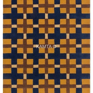 Puede incluir: Una alfombra rectangular con un patrón geométrico. La alfombra presenta un fondo azul oscuro con un patrón repetitivo de cuadrados amarillo mostaza y cruces marrones. El texto "KAMTA ©" es visible en el centro.