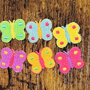 May include: Six colorful butterfly patches in shades of green, blue, yellow, pink, and purple. Each butterfly has a different color combination with embroidered details and a textured appearance. The patches are arranged on a wooden surface.