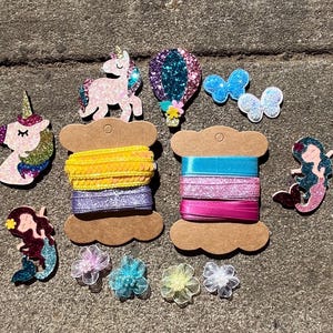 May include: A collection of colorful craft supplies and embellishments, including glittery unicorn and mermaid shapes, butterfly and flower accents, and spools of yellow, purple, blue, and pink ribbon. Perfect for DIY projects and crafting.