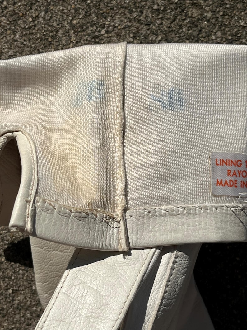 May include: Close-up of a white fencing glove, showing the interior lining and exterior leather. The glove has a stitched seam and a label with text. There are blue stains on the fabric.