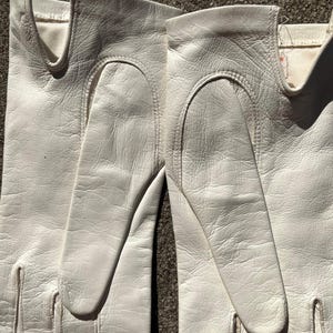 May include: A pair of white leather gloves. The gloves are made of a soft, textured leather and feature a classic design with a curved opening for the wrist. The gloves are a bright white color and appear to be in good condition.