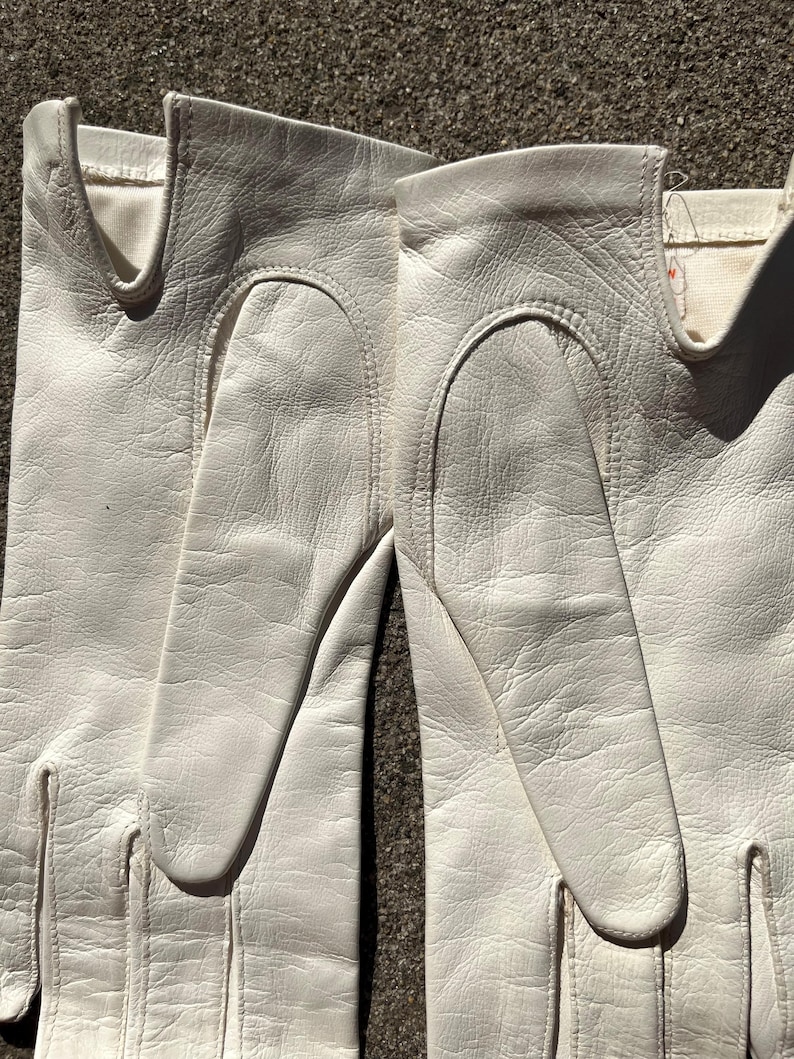 May include: A pair of white leather gloves. The gloves are made of a soft, textured leather and feature a classic design with a curved opening for the wrist. The gloves are a bright white color and appear to be in good condition.