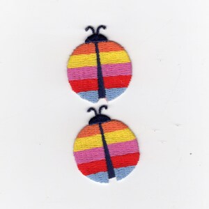 May include: Two embroidered ladybug patches. Each patch features a colorful design with horizontal stripes in red, pink, yellow, and blue. A dark blue vertical line runs down the center of each ladybug, with dark blue antennae.
