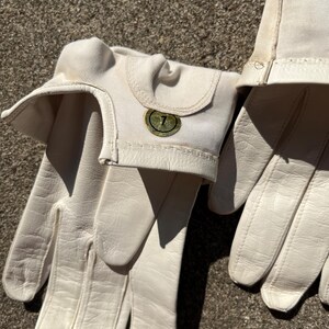 May include: A pair of off-white leather gloves with a canvas cuff. The gloves have a textured leather palm and fingers. A small, circular emblem with the number 7 is on the cuff.