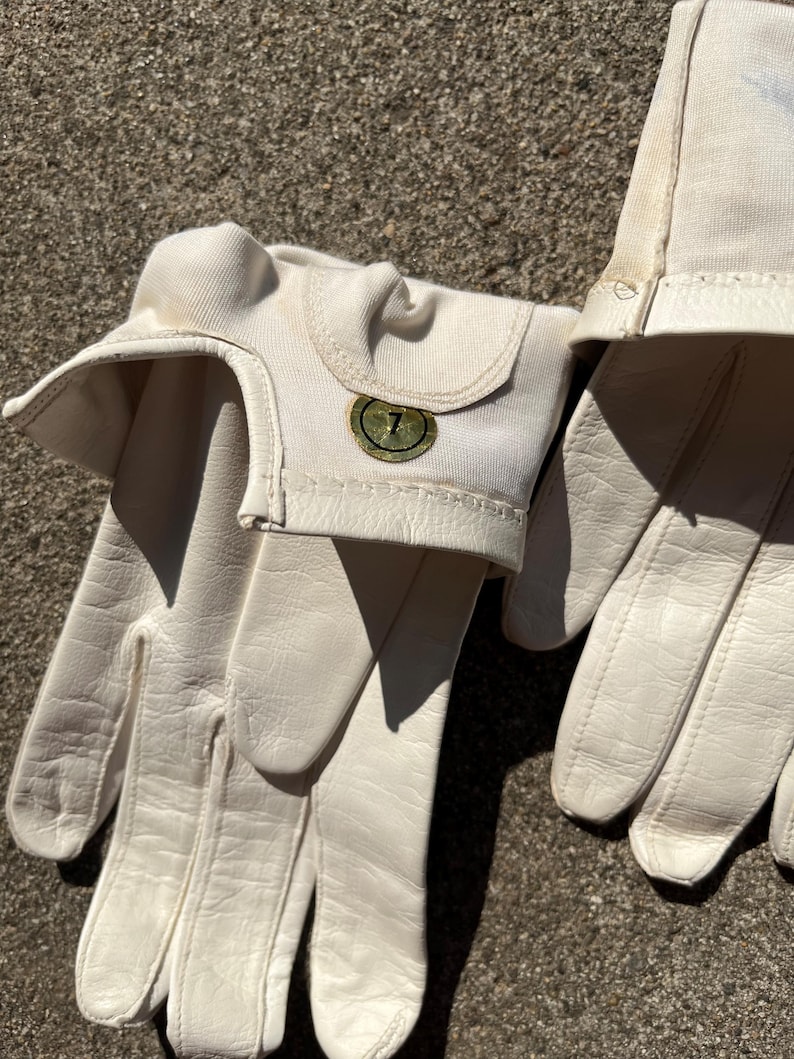 May include: A pair of off-white leather gloves with a canvas cuff. The gloves have a textured leather palm and fingers. A small, circular emblem with the number 7 is on the cuff.