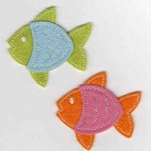 May include: Two felt fish decorations. One fish is green with a blue accent, and the other is orange with a pink accent. Both fish have white stitched details and are set against a white background.
