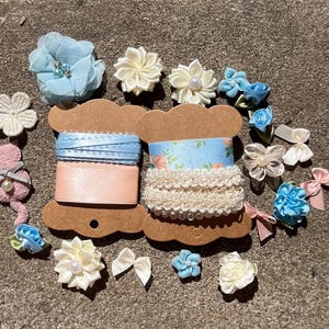 May include: An assortment of craft supplies, including spools of ribbon in light blue and peach, and floral embellishments in various shades of blue, white, and pink. There are also small bows and lace trim, perfect for DIY projects.
