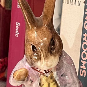 May include: A brown and white ceramic figurine of Peter Rabbit wearing a purple jacket and yellow waistcoat. He is holding a carrot in his right hand and has a mischievous expression on his face.