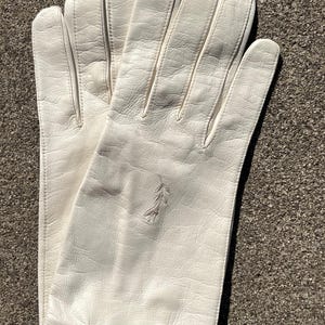 May include: A pair of off-white leather gloves. The gloves have a classic design with visible stitching and a textured surface. An embroidered floral design is visible on one glove. The gloves are likely for formal wear.
