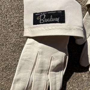 May include: A pair of white leather gloves with a cuff. The cuff has a black label that reads "MADE IN WEST GERMANY FOR the Broadway" in white script. The gloves are made of a smooth, textured leather.