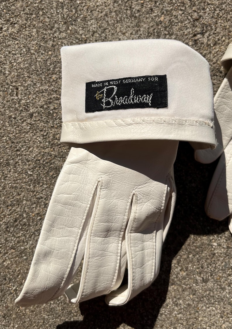 May include: A pair of white leather gloves with a cuff. The cuff has a black label that reads "MADE IN WEST GERMANY FOR the Broadway" in white script. The gloves are made of a smooth, textured leather.