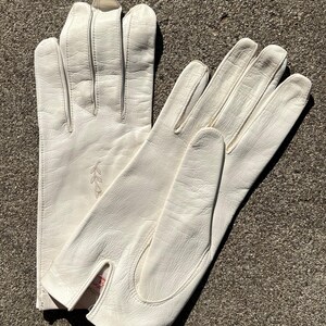 May include: A pair of off-white leather gloves. The gloves have a classic design with a split at the wrist and decorative stitching. A small embroidered floral design is visible on one glove. The gloves are laid out on a textured surface.