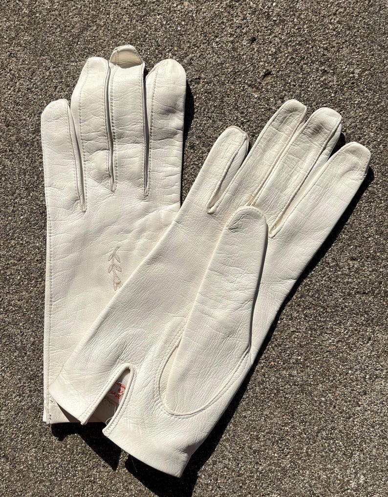 May include: A pair of off-white leather gloves. The gloves have a classic design with a split at the wrist and decorative stitching. A small embroidered floral design is visible on one glove. The gloves are laid out on a textured surface.