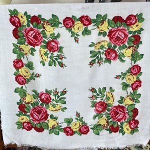 May include: A white square tablecloth with a floral design. The design features red and yellow roses with green leaves arranged in a border around the edges. The tablecloth has a fringed edge.
