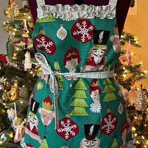 May include: A green Christmas-themed apron with a white ruffle trim. The apron features a Nutcracker design with ornaments, snowflakes, and the text "The Nutcracker".