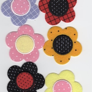 Op de afbeelding: Een verzameling van zes vilten bloemversieringen in verschillende kleuren. De bloemen hebben een bloemblaadjesontwerp met een contrasterend midden. Kleuren zijn onder meer lavendel, rood, roze, geel, zwart en oranje. De centra hebben stippen- of rasterpatronen.