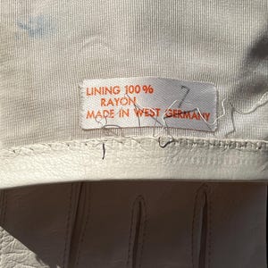 May include: Close-up of a pair of off-white gloves. The gloves have a stitched design and are made of a smooth material. A tag reads "LINING 100% RAYON MADE IN WEST GERMANY". The gloves are likely vintage.