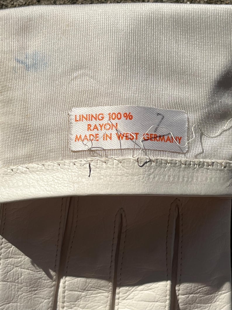May include: Close-up of a pair of off-white gloves. The gloves have a stitched design and are made of a smooth material. A tag reads "LINING 100% RAYON MADE IN WEST GERMANY". The gloves are likely vintage.
