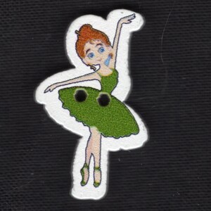 Puede incluir: Botón de madera con una bailarina de dibujos animados con un vestido verde y zapatillas de ballet. La bailarina tiene el pelo rojo y los ojos azules, con los brazos extendidos. El botón está delineado en blanco.