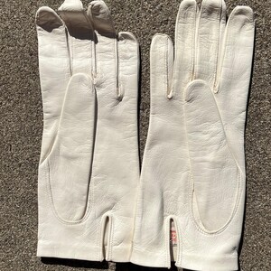 May include: A pair of off-white leather gloves. The gloves have a classic design with a split at the wrist and are made of a smooth, textured material. The gloves are laid out on a textured surface.