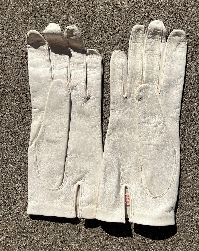 May include: A pair of off-white leather gloves. The gloves have a classic design with a split at the wrist and are made of a smooth, textured material. The gloves are laid out on a textured surface.